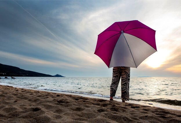 A woman stands behind a Schneider Easy sunshade in pink/white, looking out at the sea.