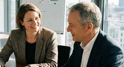 A woman and a man sit next to each other at a table in a meeting room and talk in a friendly atmosphere. AI-generated image.