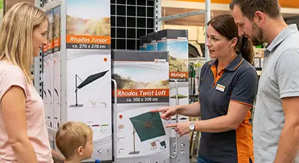 Family with their son in front of a Schneider Schirme display featuring various Rhodos models in a DIY store, while being assisted by an employee. AI-generated image.