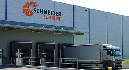 Gebäude des Schneider Schirme Hochregallagers mit drei Lkw-Laderampen, einem weißen Lkw an einer der Rampen und dem Schneider Schirme Logo an der hellgrauen Fassade. KI-generiertes BIld.