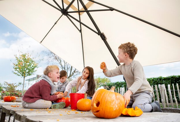 Vier Kinder sitzen unter dem Ampelschirm Rhodos Twist und schnitzen Kürbisse. Der Schirm bietet dank dreidimensionaler Verstellbarkeit optimalen UV-Schutz – ideal für Garten und Terrasse.