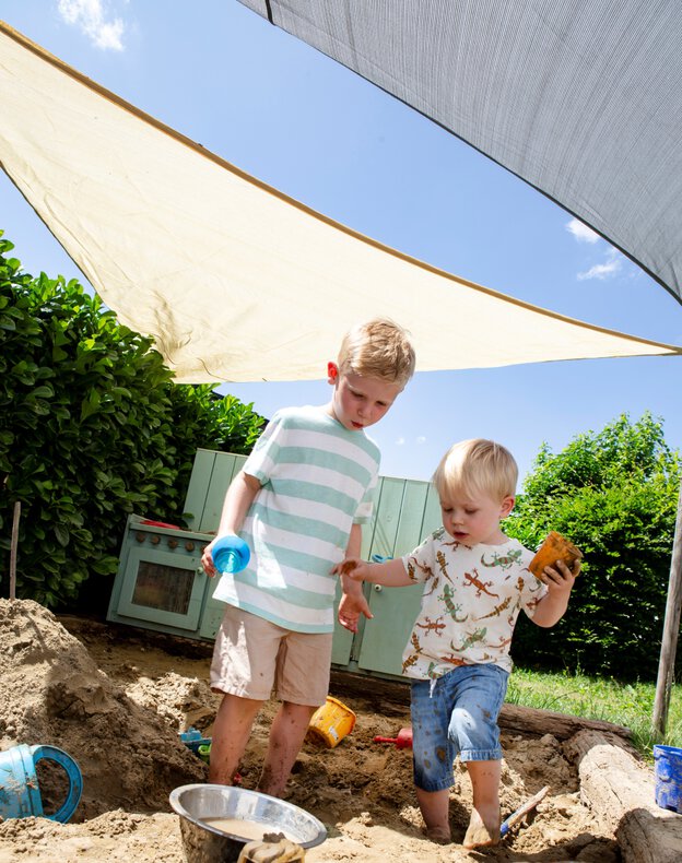 Zwei Kinder spielen im Sandkasten unter Schneider Sonnensegeln. Im Hintergrund eine Matschküche. Die Sonnensegel bieten je nach Material (HDPE oder Polyester) bis zu 90 % UV-Schutz und sind in verschiedenen Farben erhältlich.