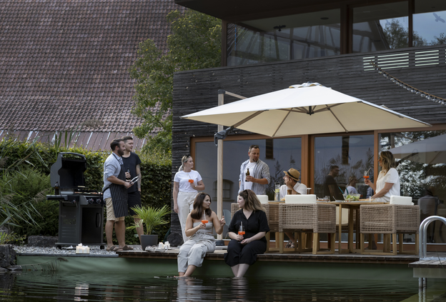 Barbecue party on the terrace with a natural pool in the foreground, where two people are dipping their feet. In the background, the Rhodos Twist Woody parasol in natural color dominates, under which people are sitting and standing with drinks.