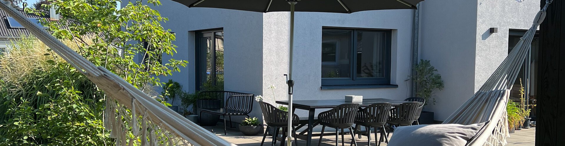 In the foreground of the image hangs a hammock, in the background, a Schneider parasol of the Monaco model stands on the terrace in front of the house. A table with chairs completes the cozy garden scene – ideal for relaxing hours in the shade.