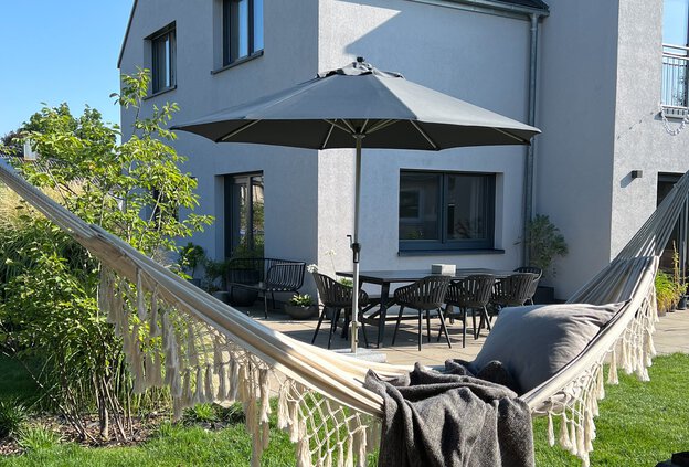 In the foreground of the image hangs a hammock, in the background, a Schneider parasol of the Monaco model stands on the terrace in front of the house. A table with chairs completes the cozy garden scene – ideal for relaxing hours in the shade.