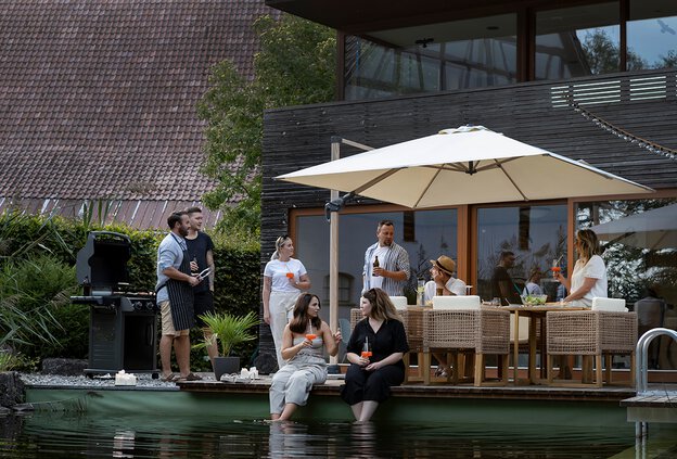Barbecue party on the terrace with a natural pool in the foreground, where two people are dipping their feet. In the background, the Rhodos Twist Woody sunshade in natural color dominates, under which people are sitting and standing with drinks.