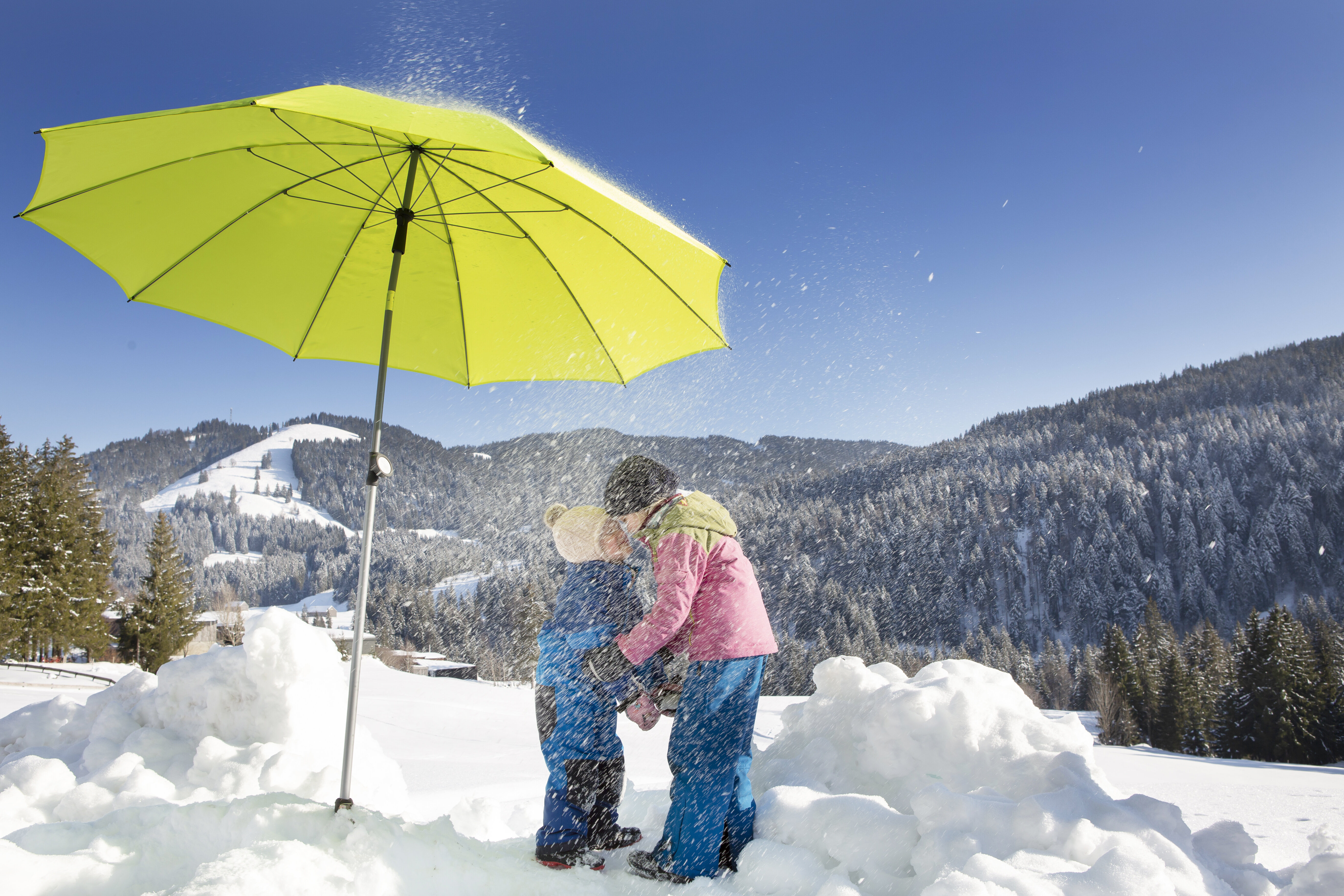 Kinder stehen im Schatten eines apfelgrünen Schneider Sonnenschirms Locarno, vor winterlichem Waldhintergrund. Der Schirm bietet effektiven Sonnenschutz, auch im Winter.