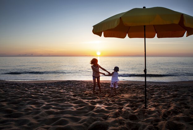 Zwei Kinder spielen am Meer bei Sonnenuntergang, daneben der goldgelbe Sonnenschirm Aquila mit Volant und neigbarem Dach – ideal bei tiefstehender Sonne, UV-Schutz 80 inklusive.