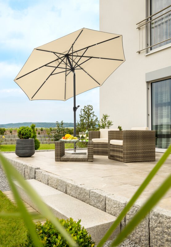 Schneider Adria parasol in ecru, 300 cm in diameter, on a terrace with lounge furniture underneath. A fruit bowl, a bottle and glasses are on the table. A stepped retaining wall made of concrete blocks is in the background. The terrace is surrounded by grass and shrubs.