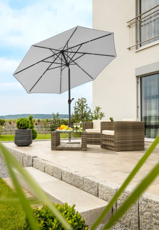 Schneider Adria parasol in silver grey, 300 cm in diameter, on a terrace with lounge furniture underneath. On the table is a fruit bowl, a bottle and glasses. In the background, a stepped retaining wall made of concrete blocks. The terrace is surrounded by grass and shrubs.