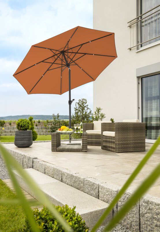Schneider Adria parasol in terracotta, 300 cm in diameter, on a terrace with lounge furniture underneath. On the table is a fruit bowl, a bottle and glasses. In the background, a stepped retaining wall made of concrete blocks. The terrace is surrounded by grass and shrubs.