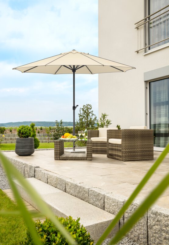 Schneider Adria parasol in ecru finish, 350 cm in diameter, on a terrace with lounge furniture underneath. A fruit bowl, a bottle and glasses are on the table. A stepped retaining wall made of concrete blocks is in the background. The terrace is surrounded by grass and shrubs.