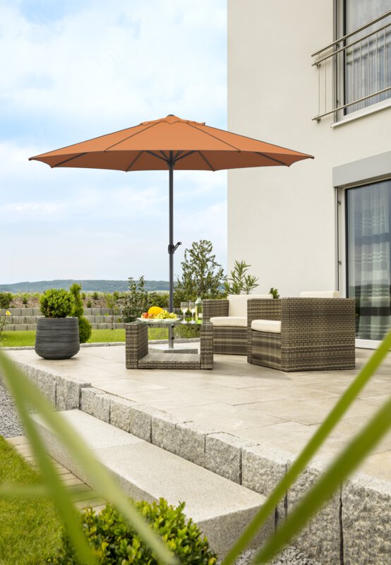 Schneider Adria parasol in terracotta, 350 cm in diameter, on a terrace with lounge furniture below. A fruit bowl, a bottle and glasses are on the table. In the background, a stepped retaining wall made of concrete blocks. The terrace is surrounded by grass and shrubs.