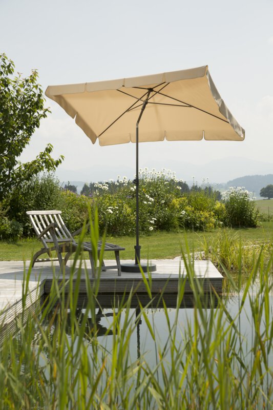 Schneider Aquila parasol in ecru, 225x120 cm, on a wooden terrace by a natural swimming pool. A deck chair sits beneath it. Reeds are in the foreground and meadows and shrubs are in the background.