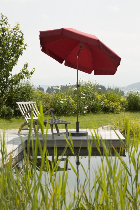 A dark red Schneider Aquila parasol, 200 cm in diameter, on a wooden deck by a natural swimming pool. A deck chair sits beneath it. Reeds are in the foregroun and meadows and shrubs are in the background.