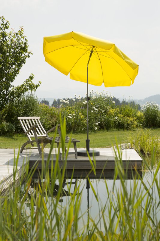 A golden-yellow Schneider Aquila parasol, 200 cm in diameter, on a wooden deck by a natural swimming pool. A deck chair sits beneath it. Reeds are in the foreground and meadows and shrubs are in the background.
