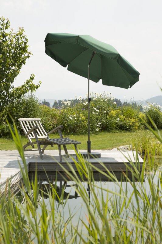 Schneider Aquila parasol in green, 200 cm in diameter, on a wooden terrace by a natural swimming pool. A deck chair sits beneath it. Reeds are in the foreground and meadows and shrubs are in the background.