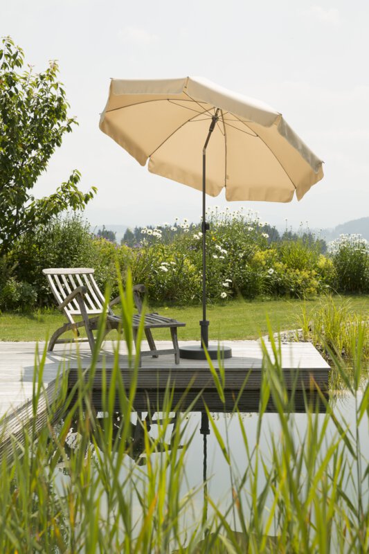 Schneider Aquila parasol in ecru, 200 cm in diameter, on a wooden terrace by a natural swimming pool. A deck chair sits beneath it. Reeds are in the foreground and meadows and shrubs are in the background.