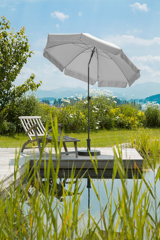Schneider Aquila parasol in silver grey, 200 cm in diameter, on a wooden terrace by a natural swimming pool. A deck chair sits beneath it. Reeds are in the foreground and meadows and shrubs are in the background.