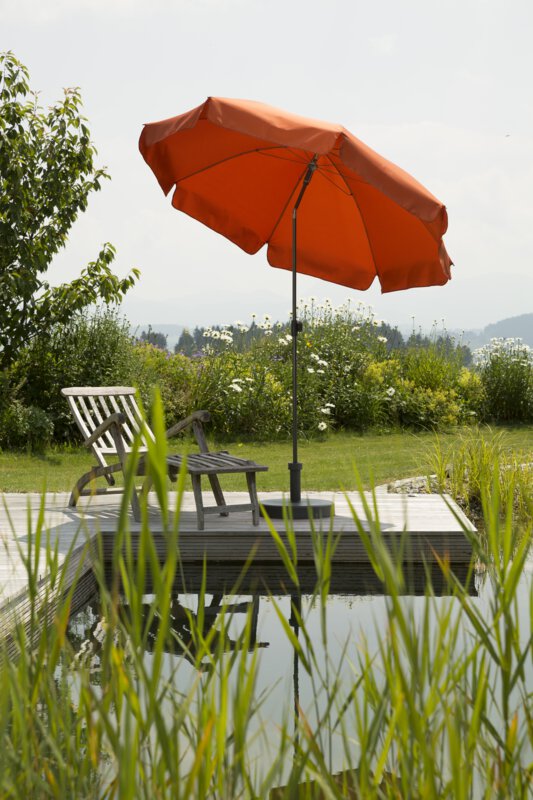 Schneider Aquila terracotta parasol, 200 cm in diameter, on a wooden terrace by a natural swimming pool. A deck chair sits beneath it. Reeds are in the foreground and meadows and shrubs are in the background.