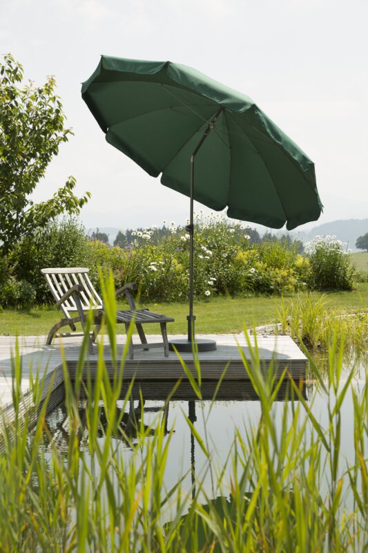 Schneider Aquila parasol in green, 250 cm in diameter, on a wooden terrace by a natural swimming pool. A deck chair sits beneath it. Reeds are in the foreground and meadows and shrubs are in the background.