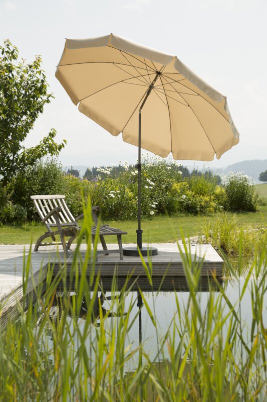 Schneider Aquila parasol in ecru, 250 cm in diameter, on a wooden terrace by a natural swimming pool. A deck chair sits beneath it. Reeds are in the foreground and meadows and shrubs are in the background.