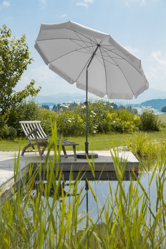 Schneider Aquila parasol in silver grey, 250 cm in diameter, on a wooden terrace by a natural swimming pool. A deck chair sits beneath it. Reeds are in the foregroun and meadows and shrubs are in the background.