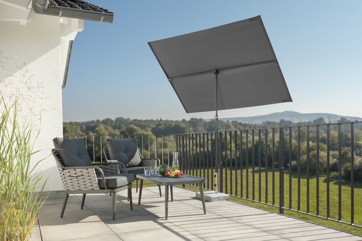 Schneider Sonnenschirm Avellino in Anthrazit, 180x130 cm, auf einer Dachterrasse. Im Schatten des Schirms stehen zwei Stühle mit grauen Sitzpolstern und ein kleiner Tisch mit Obstschale, Karaffe und Gläsern. Im Hintergrund Wiese, Wald und ein Hügel unter strahlend blauem Himmel.