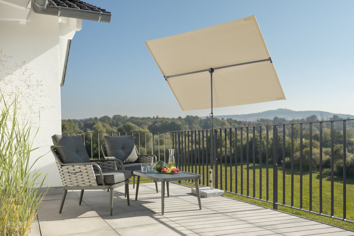 Schneider Sonnenschirm Avellino in Natur, 180x130 cm, auf einer Dachterrasse. Im Schatten des Schirms stehen zwei Stühle mit grauen Sitzpolstern und ein kleiner Tisch mit Obstschale, Karaffe und Gläsern. Im Hintergrund Wiese, Wald und ein Hügel unter strahlend blauem Himmel.