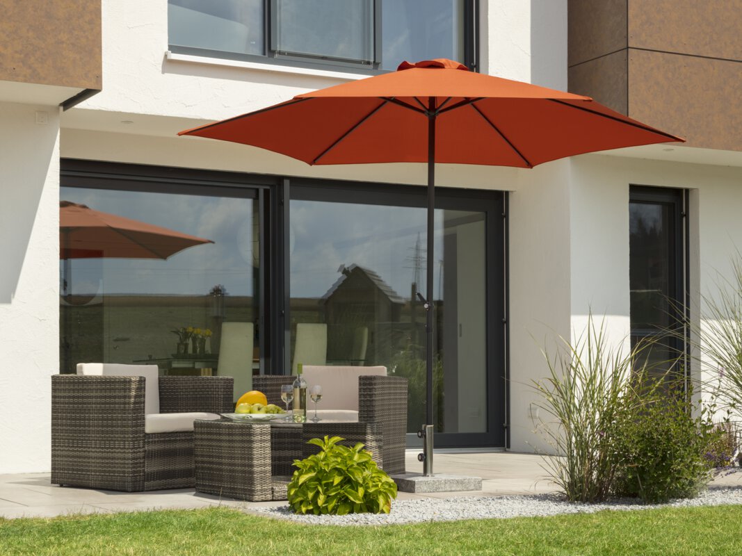 A Schneider Bergamo terracotta parasol, 300 cm in diameter, on a terrace with lounge furniture. A fruit bowl with a bottle and glasses sits on the table. Small shrubs and a meadow are in the foreground.
