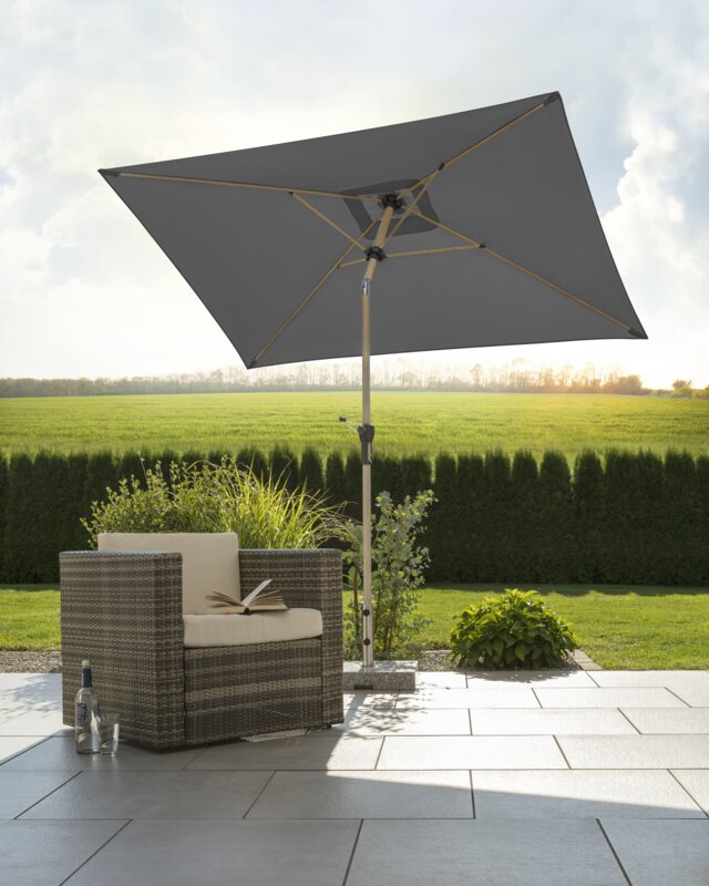 Schneider parasol Cordoba in anthracite, 230x150 cm, on a terrace with a deck chair. An open book lies on the chair and a bottle and a glass sit beside it. In the background are small shrubs, a meadow, hedges and in the distance a forest, with rays of sunlight shimmering through. The scene takes place away from the house.