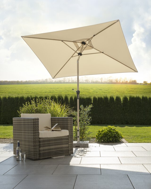 Schneider parasol Cordoba in ecru, 230x150 cm, on a terrace with a deck chair. An open book lies on the chair and a bottle and a glass sit beside it. In the background are small shrubs, a meadow, hedges and in the distance a forest, with rays of sunlight shimmering through. The scene takes place away from the house.