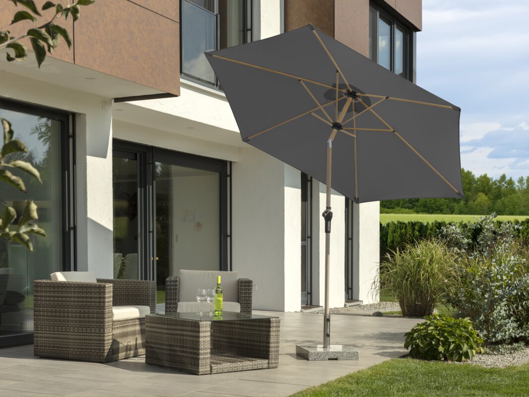 Schneider parasol Cordoba in anthracite, 270 cm in diameter, on a terrace with lounge furniture. A bottle and two glasses are on the table. In the foreground, a meadow, in the background, small shrubs, a hedge, a meadow and a forest.