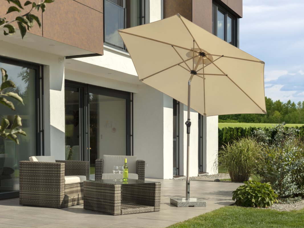 Schneider Cordoba parasol in ecru, 270 cm in diameter, on a terrace with lounge furniture. A bottle and two glasses are on the table. In the foreground, a meadow, in the background, small shrubs, a hedge, a meadow and a forest.