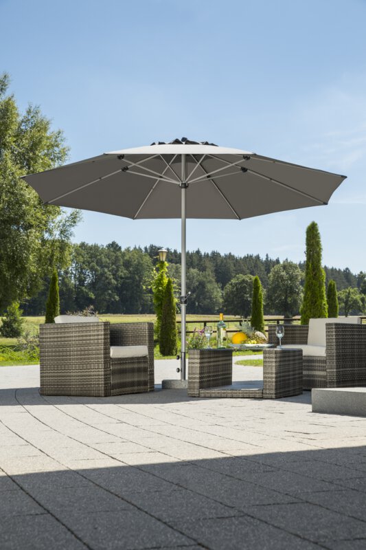 A Schneider Gemini parasol in anthracite, 360 cm in diameter, on a terrace with lounge furniture. A fruit bowl and a bottle with two glasses sit on the table. In the background are shrubs, a fence, a meadow with trees and a forest.