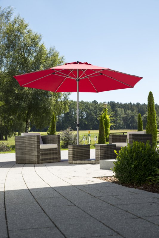 A Schneider Gemini parasol in dark red, 360 cm in diameter, on a terrace with lounge furniture. A fruit bowl and a bottle with two glasses sit on the table. In the background are shrubs, a fence, a meadow with trees and a forest.