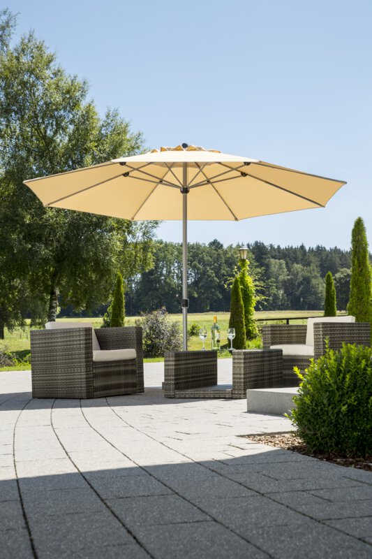 Schneider Sonnenschirm Gemini in Natur, 360 cm Durchmesser, auf einer Terrasse mit Lounge-Möbeln. Auf dem Tisch befindet sich eine Flasche mit zwei Gläsern. Im Hintergrund Sträucher, ein Zaun, eine Wiese mit Bäumen und Wald.
