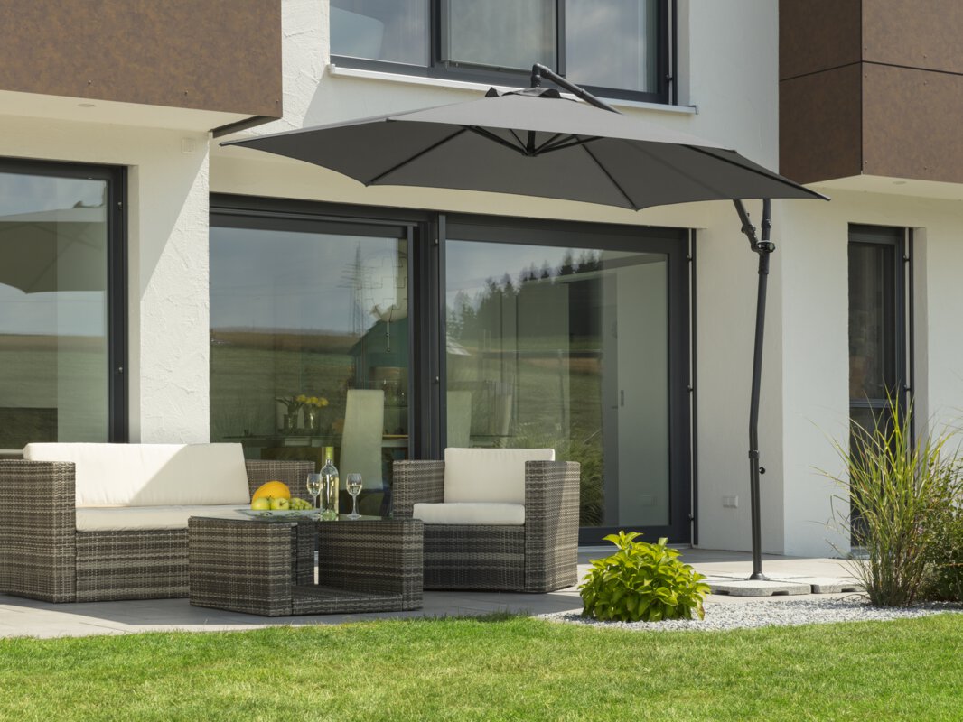 Schneider Genua parasol in anthracite, 300 cm in diameter, on a terrace with lounge furniture. On the table is a fruit bowl with a bottle and glasses. Small shrubs and a meadow are in the foreground.