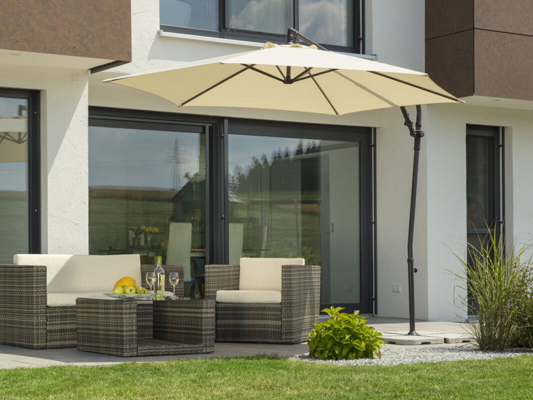A Schneider Genua parasol in ecru, 300 cm in diameter, on a terrace with lounge furniture. A fruit bowl with a bottle and glasses sits on the table. Small shrubs and a meadow are in the foreground.