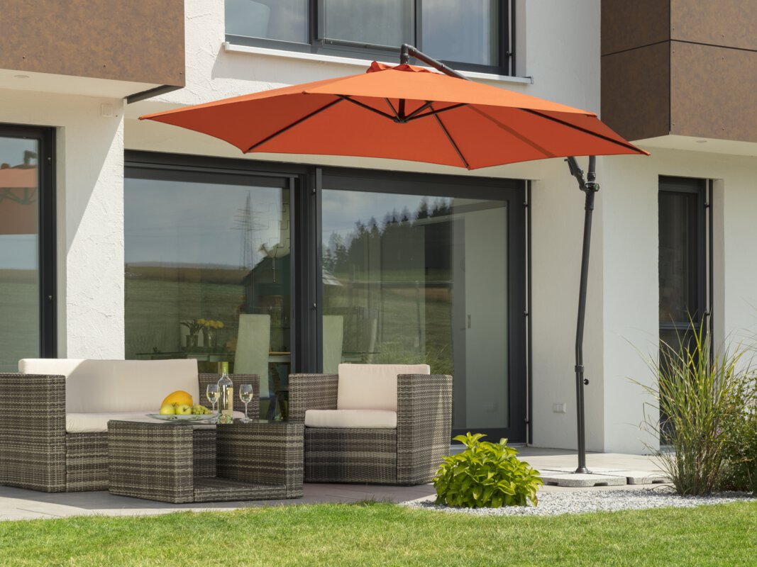 A Schneider Genua parasol in terracotta, 300 cm in diameter, on a terrace with lounge furniture. A fruit bowl with a bottle and glasses sits on the table. Small shrubs and a meadow are in the foreground.