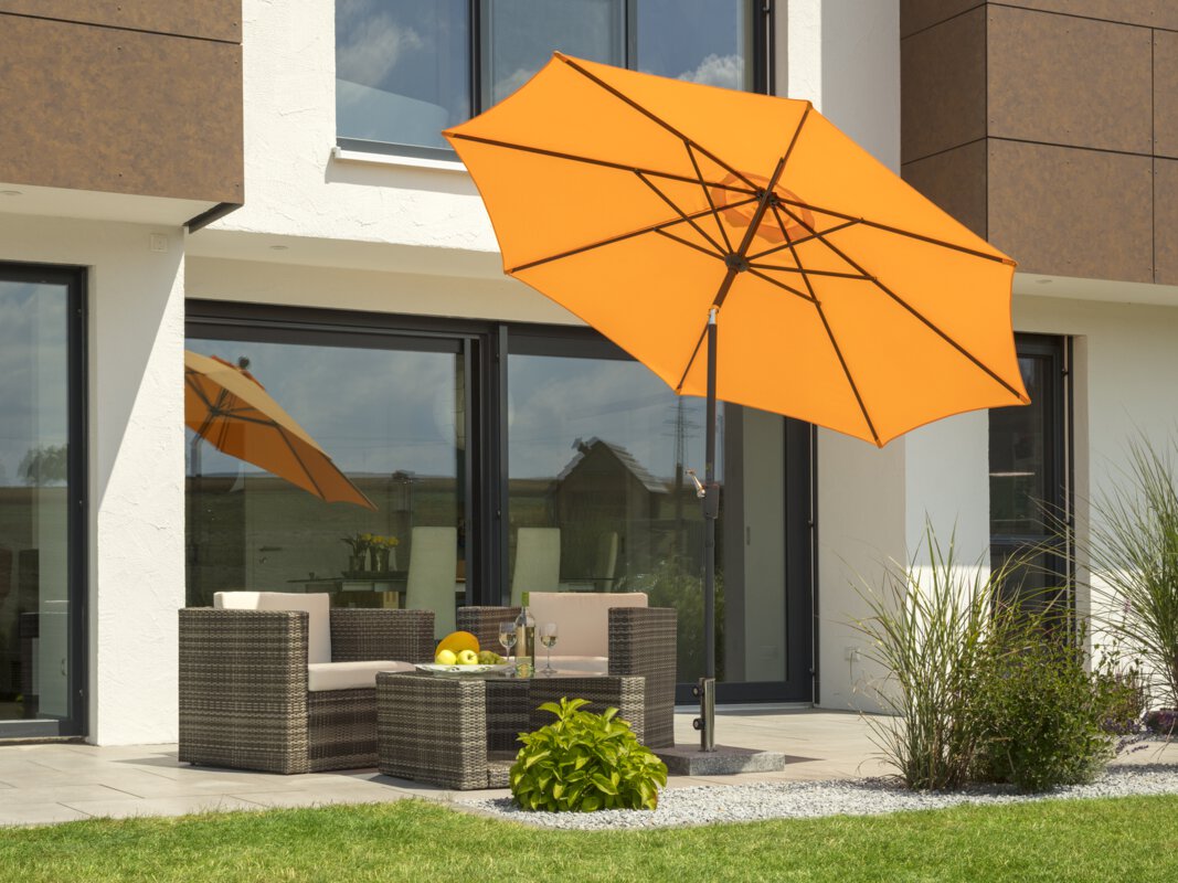 A Schneider Harlem parasol in mandarin, 270 cm in diameter, is on a terrace with lounge furniture. A fruit bowl with a bottle and two glasses sits on the table. Small shrubs and a lawn are in the foreground.