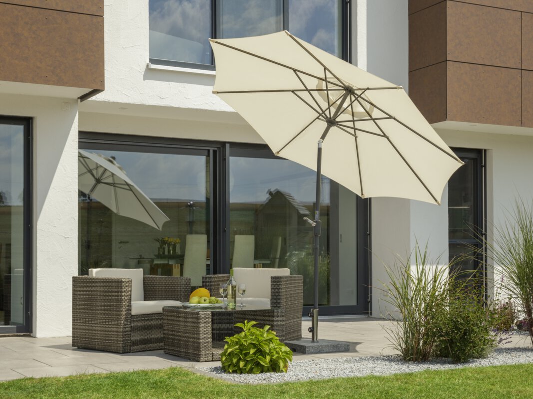 A Schneider Harlem parasol in ecru, 270 cm in diameter, on a terrace with lounge furniture. A fruit bowl with a bottle and two glasses sits on the table. Small shrubs and a lawn are in the foreground.