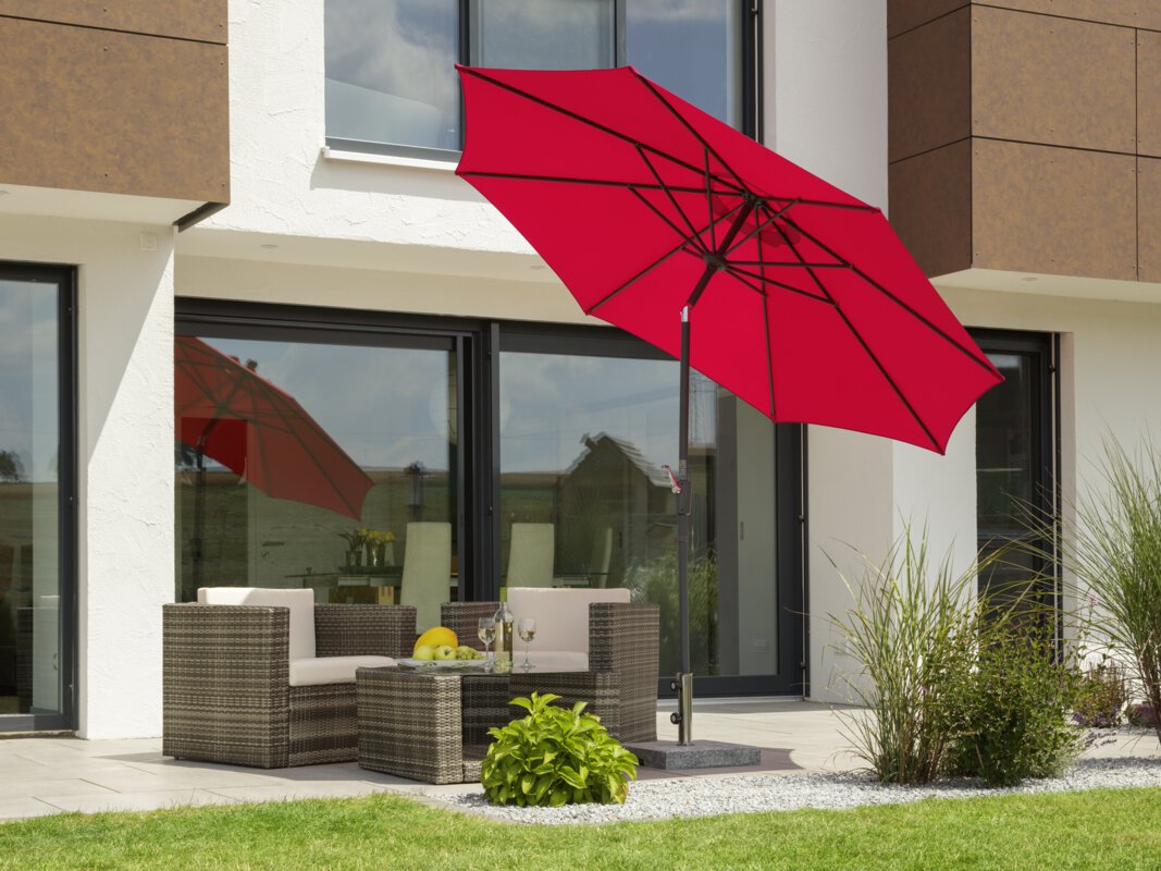 A Schneider Harlem parasol in red, 270 cm in diameter, on a terrace with lounge furniture. A fruit bowl with a bottle and two glasses sits on the table. Small shrubs and a lawn are in the foreground.