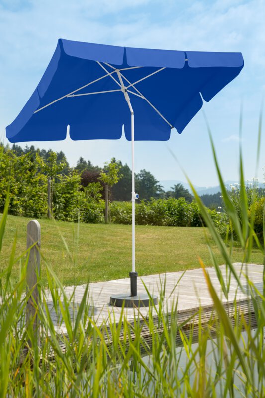 Schneider Ibiza parasol in blue, 180x120 cm, on a wooden terrace by a natural swimming pool. Reeds in the foreground, meadow and various green plants in the background.