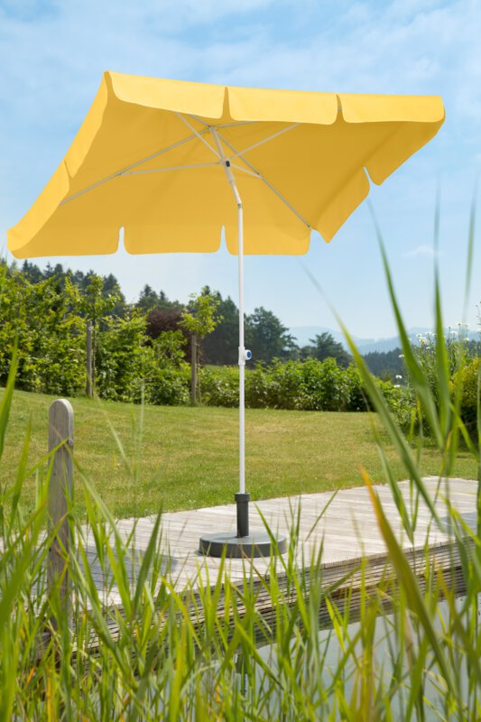 Schneider Ibiza parasol in gold yellow, 180x120 cm, on a wooden terrace by a natural swimming pool. Reeds in the foreground, meadow and various green plants in the background.