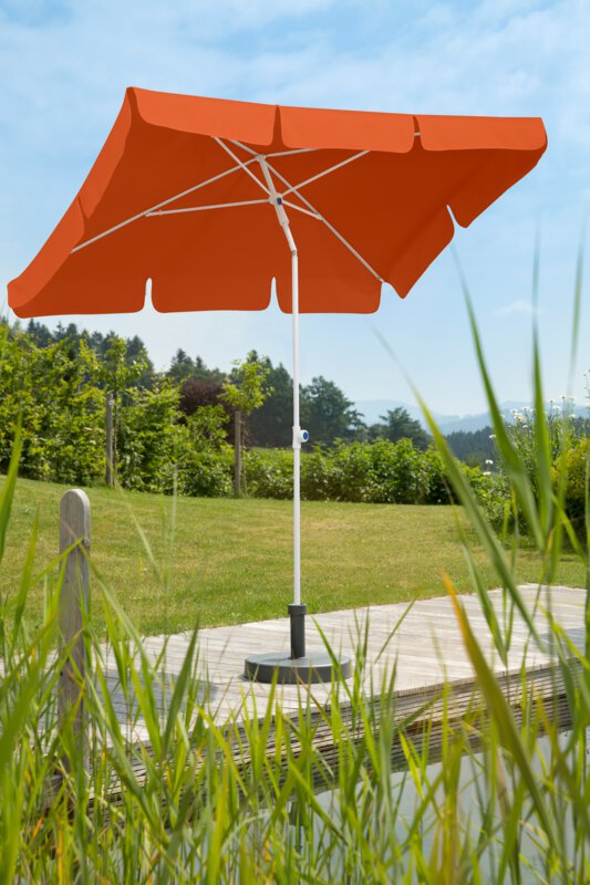 Schneider Ibiza parasol in terracotta, 180x120 cm, on a wooden terrace by a natural swimming pool. Reeds in the foreground, meadow and various green plants in the background.