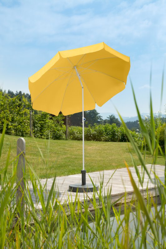 A golden-yellow Schneider Ibiza parasol, 200 cm in diameter, on a wooden terrace by a natural swimming pool. Reeds are in the foreground, meadow and various green plants are in the background.