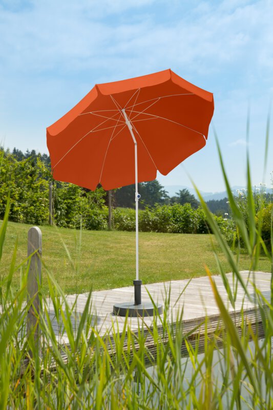 Schneider Ibiza parasol in terracotta, 200 cm in diameter, on a wooden terrace by a natural swimming pool. Reeds in the foreground, meadow and various green plants in the background.