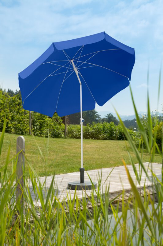 Schneider Ibiza parasol in blue, 240 cm in diameter, on a wooden terrace by a natural swimming pool. Reeds in the foreground, meadow and various green plants in the background.