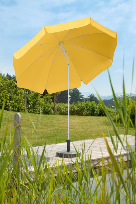A golden-yellow Schneider Ibiza parasol, 240 cm in diameter, on a wooden terrace by a natural swimming pool. Reeds are in the foreground, meadow and various green plants are in the background.